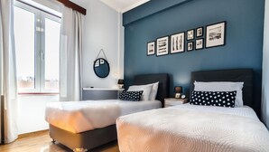 A hotel room with two beds, a large window with curtains, a wall with framed pictures, and a round mirror. - Casa Ithomi, 3 Bedroom Maisonnet, Old Harbor Chania (Χανιά)
