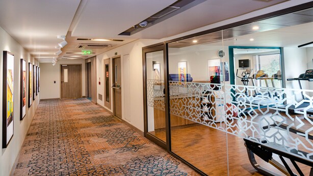 A hotel corridor with a patterned carpet, glass partitions, and exercise equipment. - Holiday Inn Express Kolkata Airport by IHG (Barakpur)
