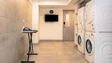 A laundry room with stacked washing machines, a flat-screen TV on the wall, and a small ironing station.