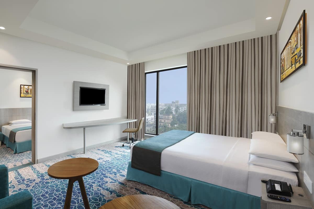 Een moderne hotelkamer met een groot bed, een aan de muur bevestigde televisie, een bureau met een stoel en uitzicht op de stad door grote ramen.