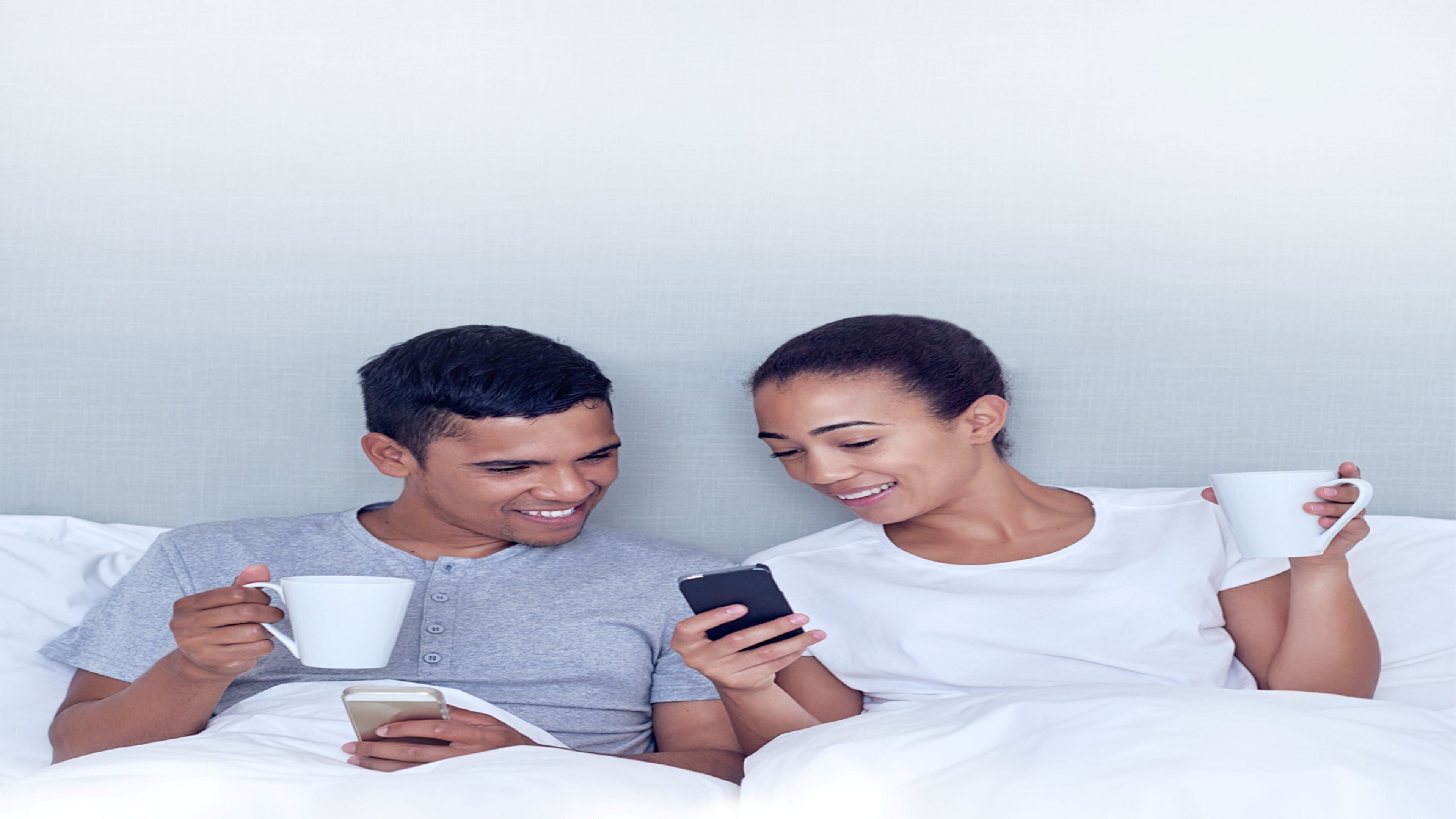 Two individuals in a bedroom with white bedding, one holding a smartphone and the other holding a white mug.