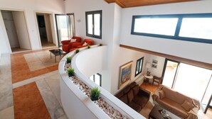 A spacious living area with a curved white bench, a brown sofa, and a red chair. There are multiple windows providing natural light, and a ceiling with wooden beams.