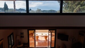 A room with a large window overlooking a terrace. The room has a bed, a small table, and a chair.