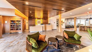 Un moderno vestíbulo de hotel con techo de madera, un mostrador de recepción, una zona de estar con sofás verdes y una cocina visible al fondo.