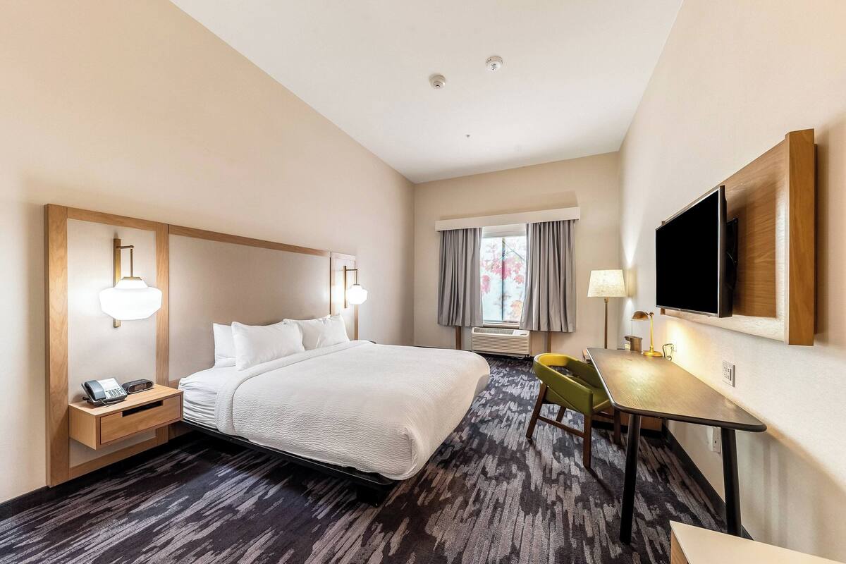Habitación de hotel con una cama grande, mesita de noche con teléfono, escritorio con silla, televisor de pantalla plana montado en la pared y ventana con cortinas.