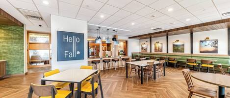 A modern restaurant with wooden flooring, yellow and brown chairs, and a 'Hello Fall' sign.