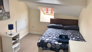 A bedroom with a bed, a desk, a lamp, a radiator, and a window with a view of buildings. - Fitzwilliam Street Rooms (Sheffield)