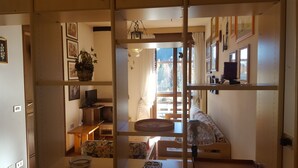 En hyggelig stue med trægulv, sofa, spisebord og et fjernsyn på et stativ. Der er reoler med pynteting og et vindue med gardiner.