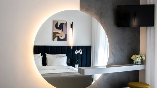 Una habitación de hotel moderna con un espejo circular, una cama con ropa de cama blanca, un cabecero oscuro, un televisor de pantalla plana montado en la pared y un pequeño florero con flores sobre una repisa.