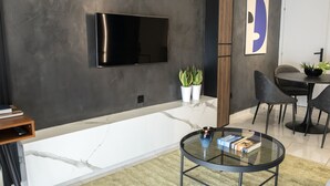 Una sala moderna con un televisor de pantalla plana montado en la pared, una mesa de centro de vidrio con libros, una zona de comedor con mesa y sillas, y plantas en macetas.
