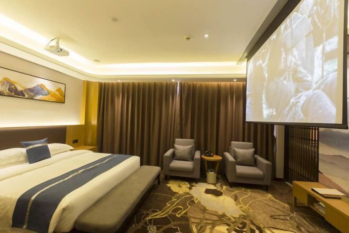 Une chambre d’hôtel comprenant un lit, deux fauteuils, une petite table, un écran de projection et une grande fenêtre avec des rideaux.