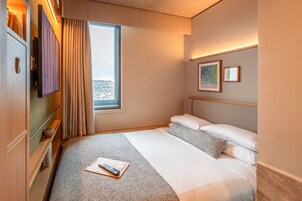 Una habitación de hotel con cama, ventana con cortinas, mesita de noche con un libro y control remoto, y una pared decorada con cuadros.