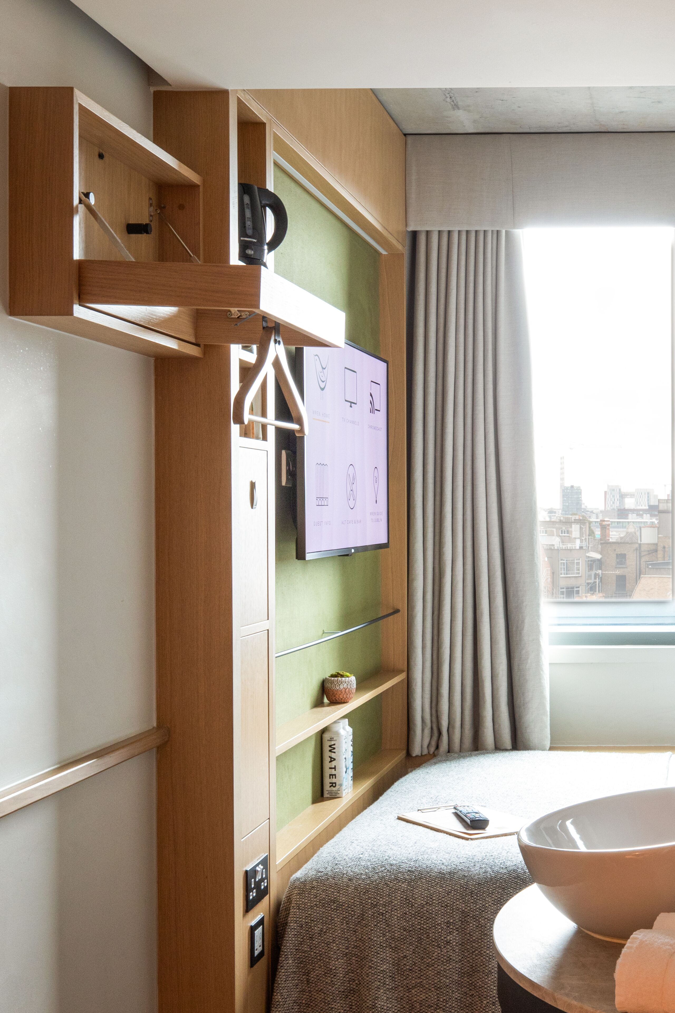 A hotel room with a wooden wardrobe, a wall-mounted TV, a large window with curtains, and a round bathtub.