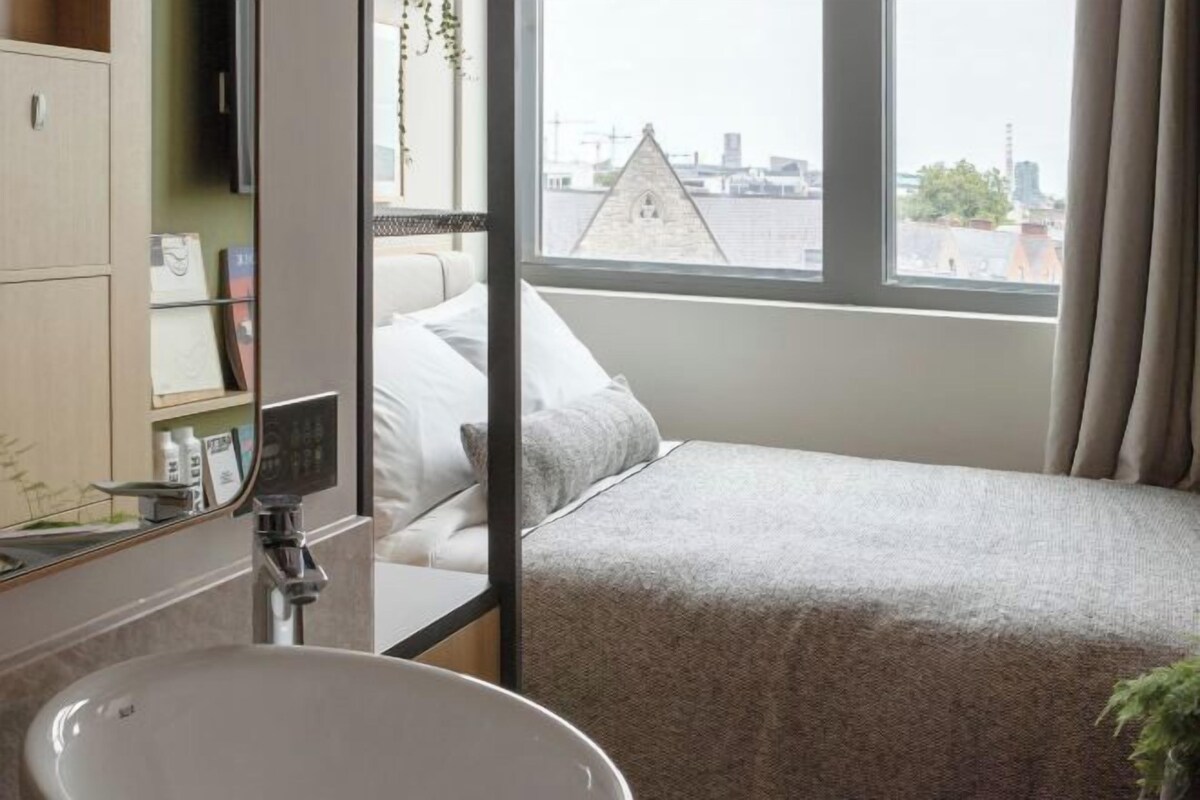 A hotel room with a bed, a window with a view of buildings, a sink, and a mirror.