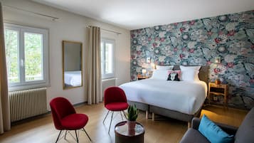 Une chambre d’hôtel avec un grand lit, deux fauteuils rouges, une table basse ronde ornée d’une plante, un canapé et un mur décoré d’un motif floral.