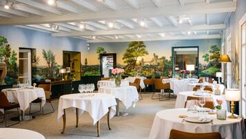 Un restaurante con mesas redondas, manteles blancos y sillas. Hay cuadros en las paredes y un ventanal grande a la derecha.