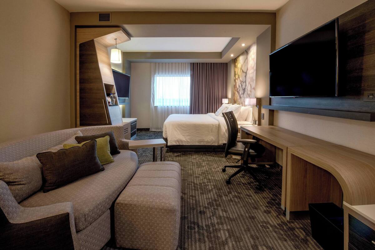 Ein Hotelzimmer mit Sofa, Schreibtisch, Bett, Fernseher und einem Fenster mit Vorhängen.