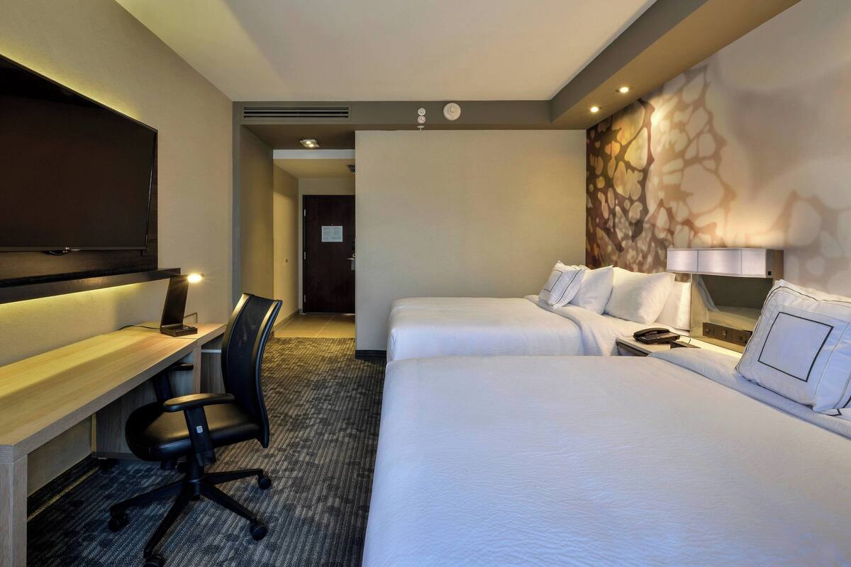 Ein Hotelzimmer mit Flachbildfernseher, Schreibtisch mit Stuhl, zwei Betten mit weißer Bettwäsche und einer Wanddekoration.