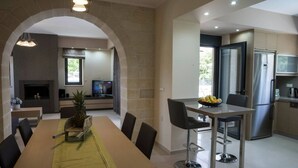 A modern kitchen with a stainless steel refrigerator, a dining area with a table and chairs, and an open-plan layout with a fireplace. - Villa 200m to Beach & Amenities w/Pool, Sauna, BBQ (Αλμυρίδα)