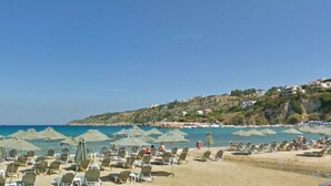 A beach with rows of sun loungers and umbrellas, a clear sky, and a hillside with buildings. - Villa 200m to Beach & Amenities w/Pool, Sauna, BBQ (Αλμυρίδα)