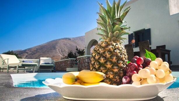 Une assiette de fruits comprenant un ananas, des bananes et des raisins, posée sur une table près d’une piscine, avec une vue sur les montagnes.