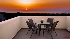 Un balcon avec une table ronde et quatre chaises, un vase rempli de fleurs jaunes et une vue sur le coucher de soleil.