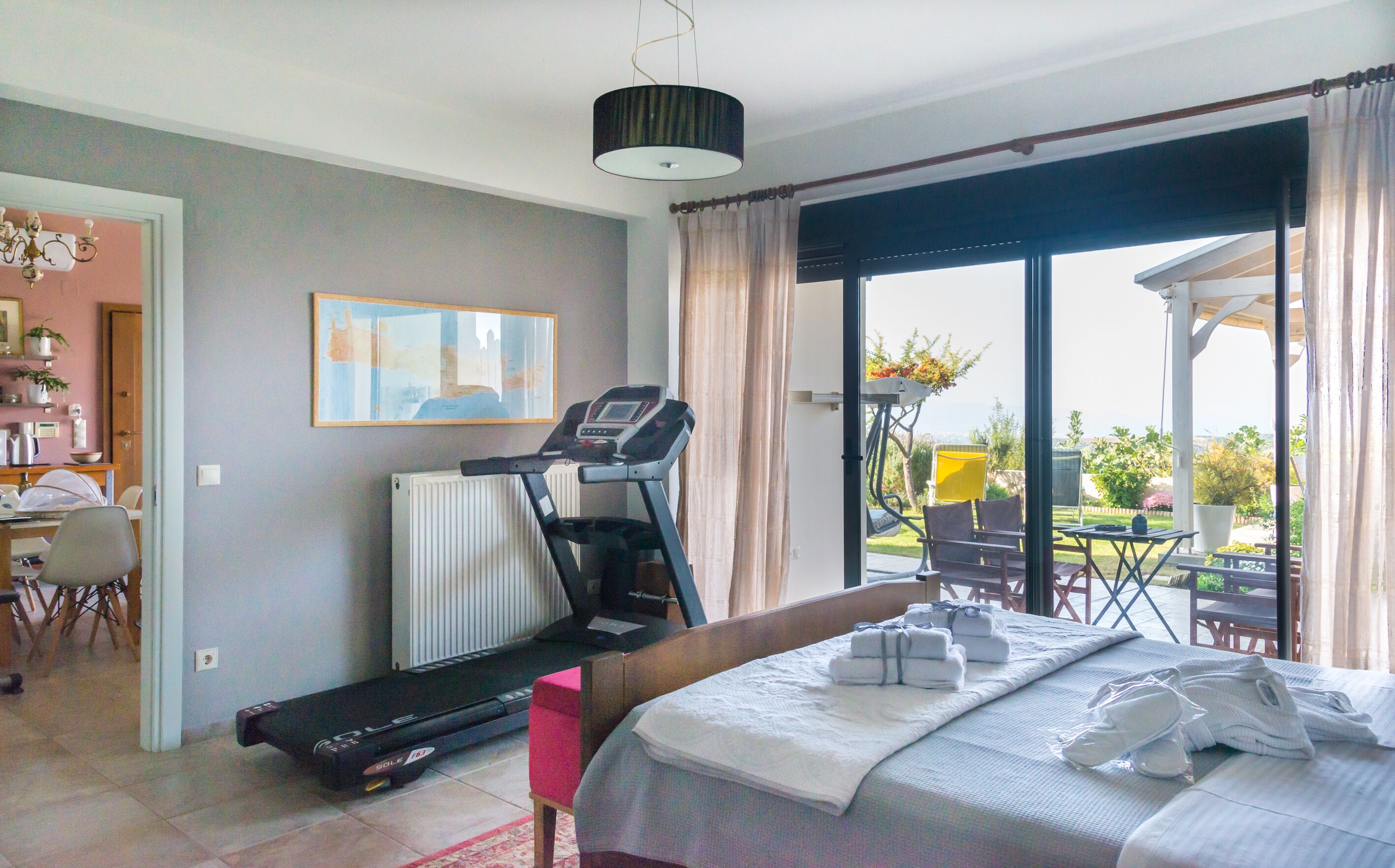 A hotel room with a treadmill, a bed with towels, a dining area, and a view of the outdoors.