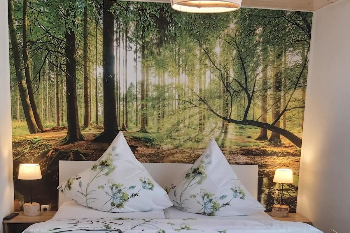 A bedroom with a large forest-themed tapestry on the wall, a bed with floral bedding, and two bedside lamps.