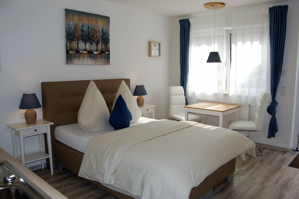 A hotel room with a bed, bedside tables, a chair, a small table, a painting, a clock, and a window with curtains.