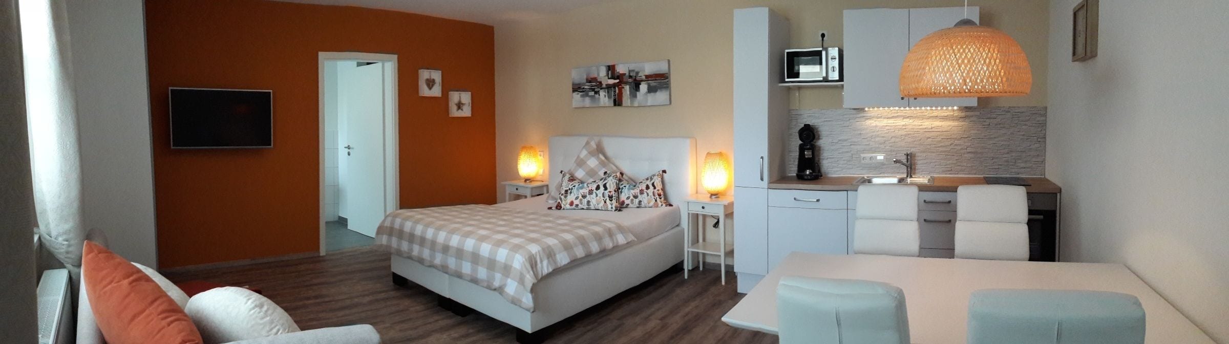 A modern hotel room with a bed, a desk, a kitchenette, and a TV.