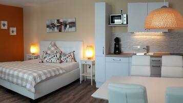 A modern hotel room with a bed, a desk, a kitchenette, and a TV.