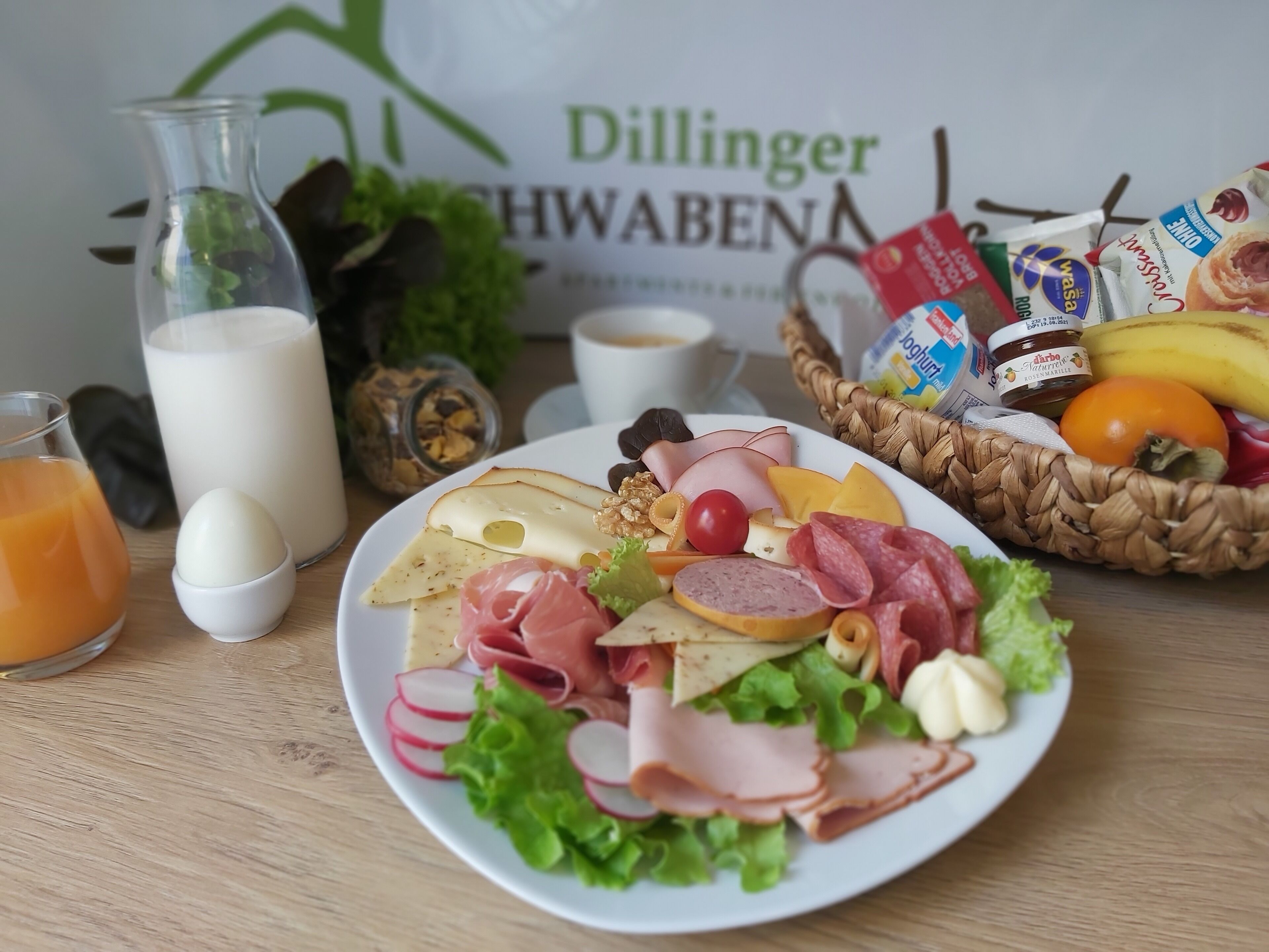 Una colazione con un piatto di salumi, formaggi e verdure, un bicchiere di succo d'arancia, un bicchiere di latte e un cesto di frutta e snack.