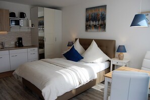 A modern hotel room with a bed, bedside tables, a desk, and a kitchenette.