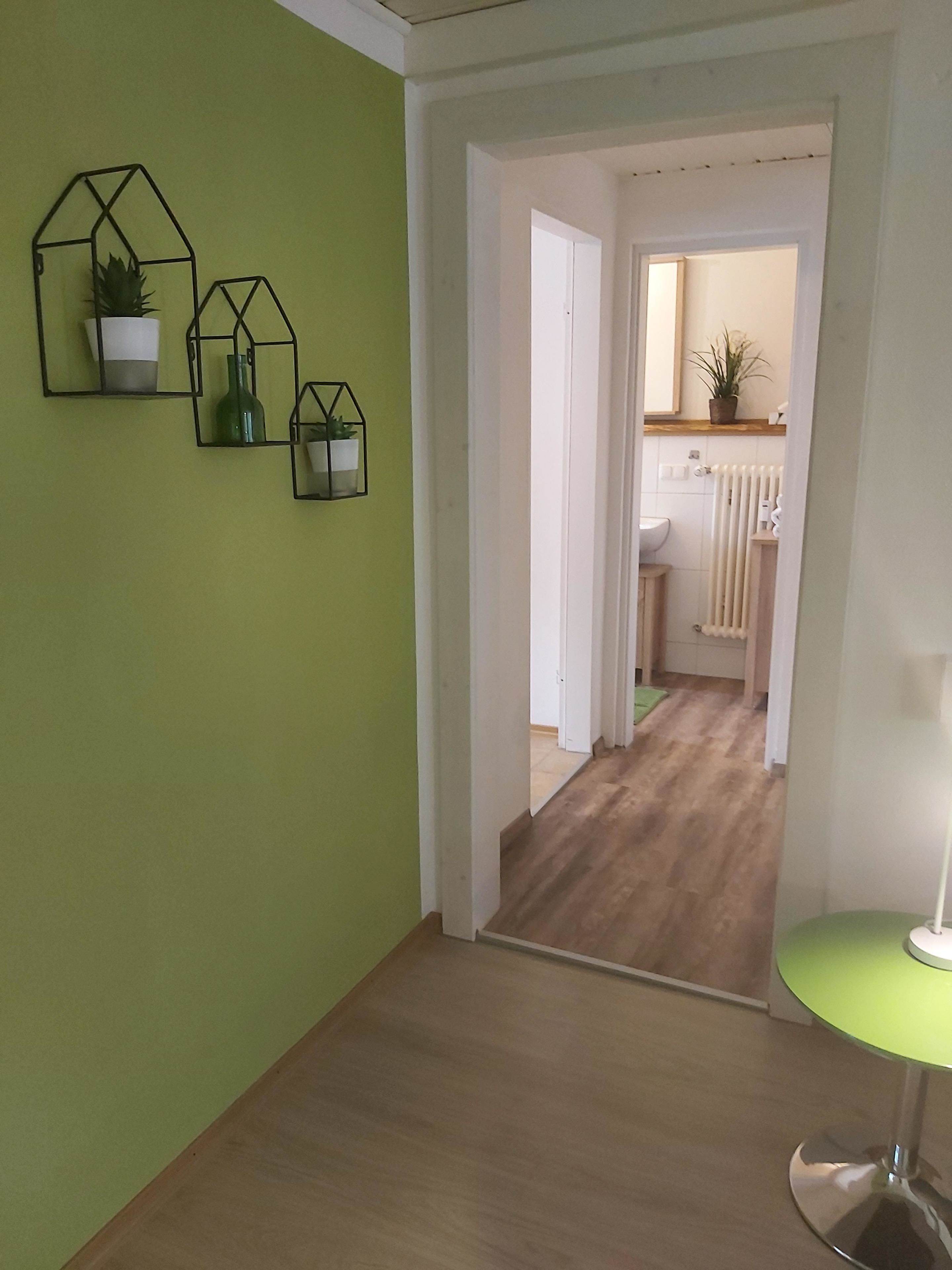A hallway with green walls, wooden flooring, and a green table lamp. There are three black geometric wall decorations with plants.