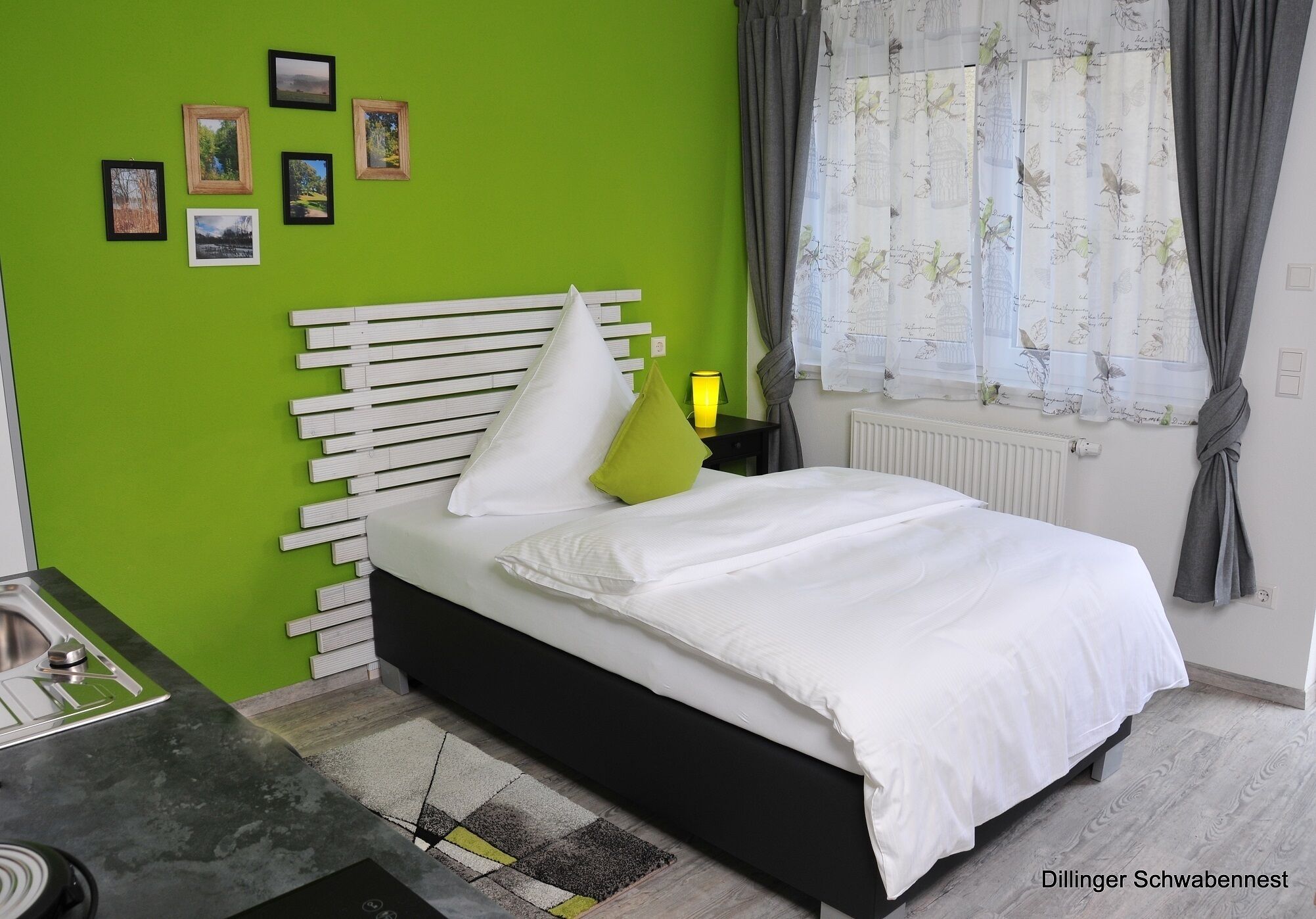 Een slaapkamer met een bed, een nachtkastje, een raam met gordijnen en een groene wand met ingelijste foto's.