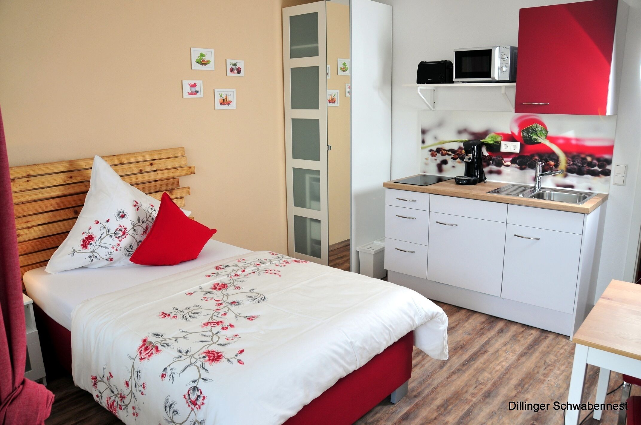 A bedroom with a wooden headboard, a bed with floral bedding, a red pillow, a small bedside table, and a kitchenette with a microwave, toaster, and coffee maker.