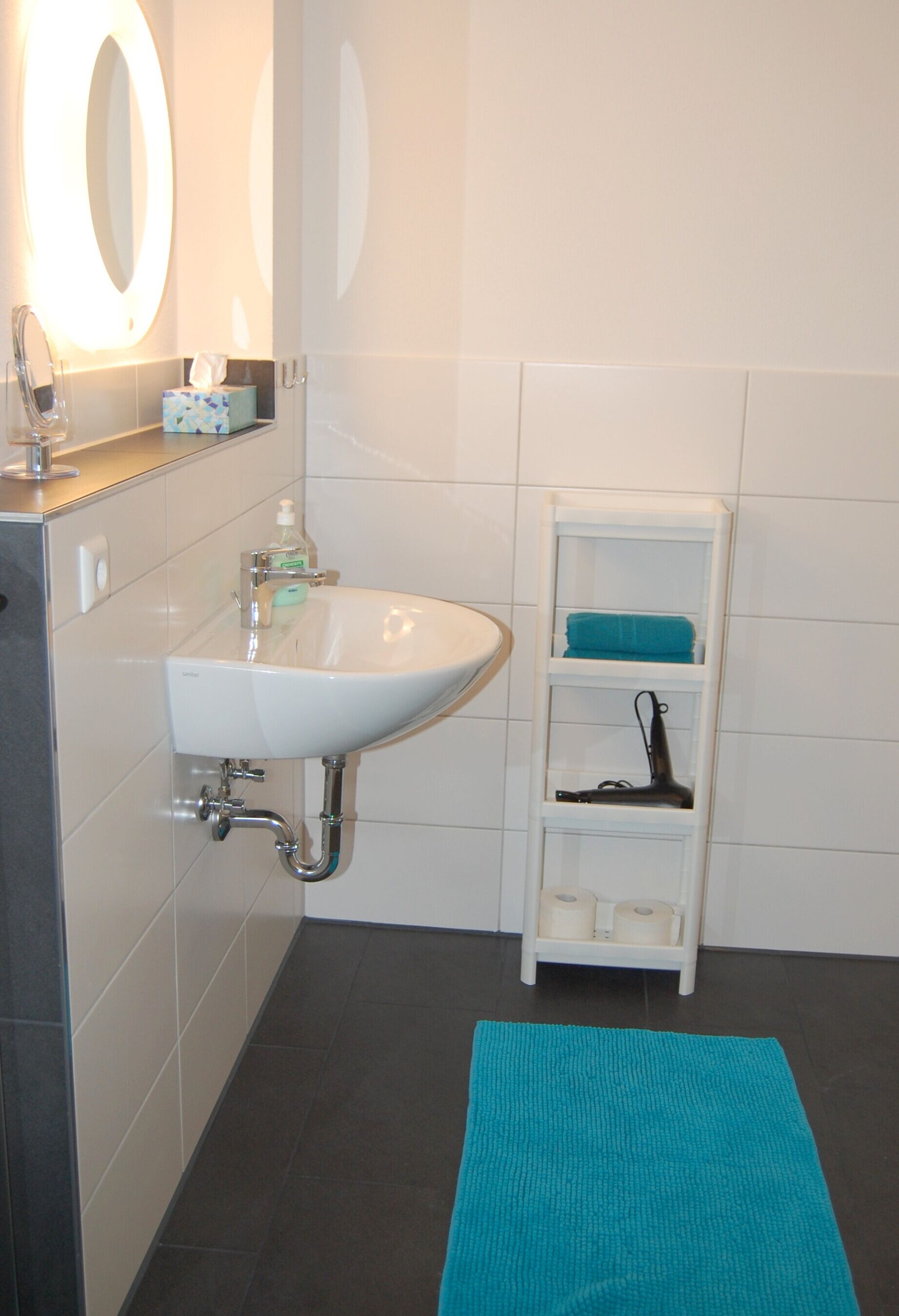 A bathroom with a white sink, a mirror with lights, a shelf with a towel, and a blue bath mat.