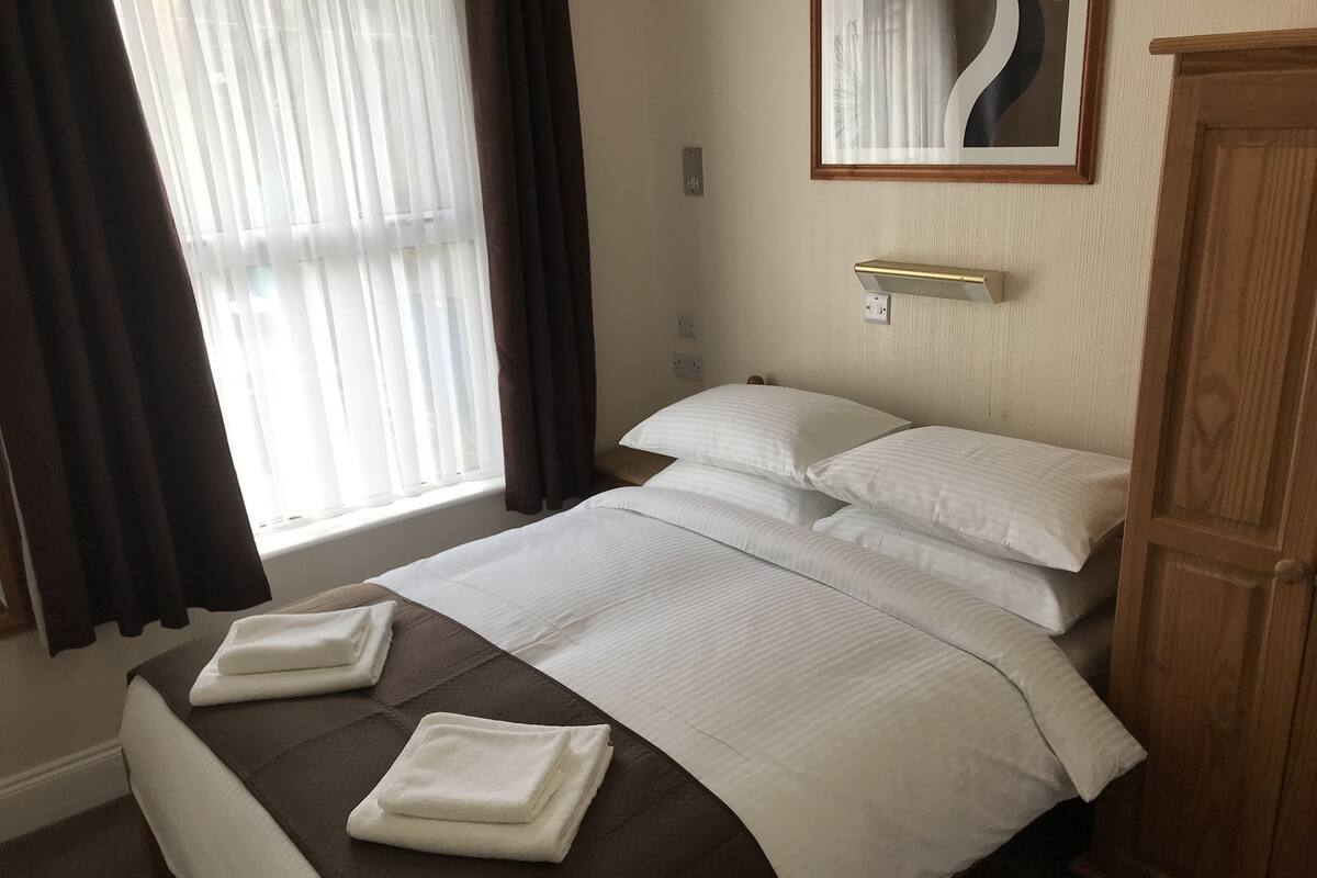 A neatly made bed with white linens and two folded towels, a wooden wardrobe, and a window with sheer curtains.