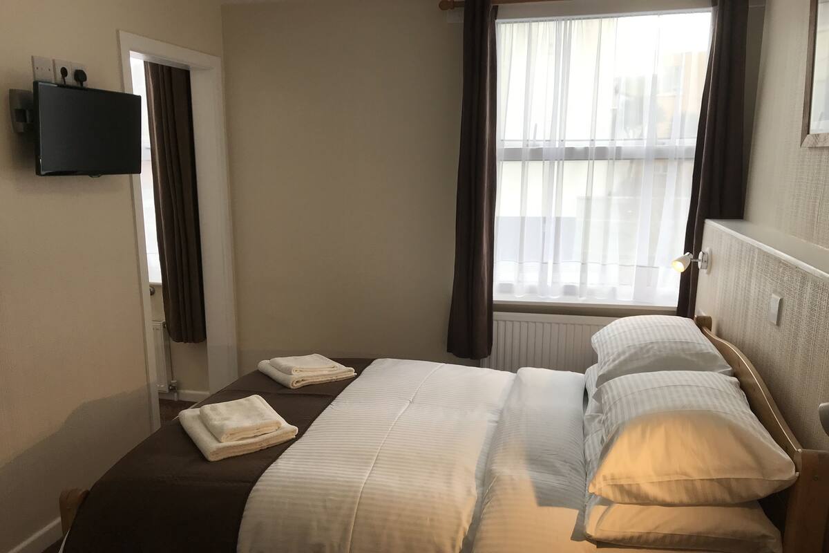 A hotel room with a bed, a TV mounted on the wall, a window with curtains, and a bedside table.