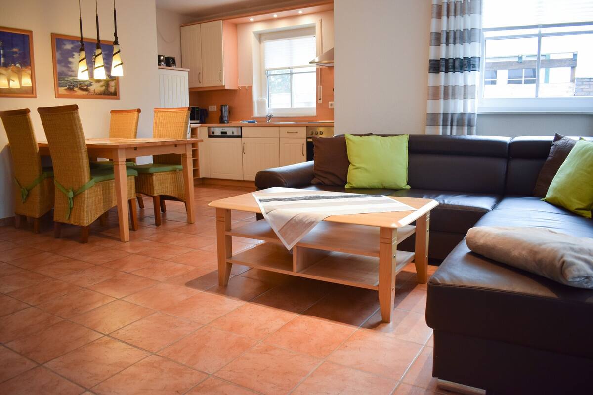 A living room with a sofa, a dining table with chairs, a kitchen area, and a window with curtains.