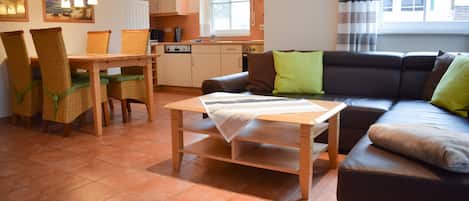 A living room with a sofa, a dining table with chairs, a kitchen area, and a window with curtains.