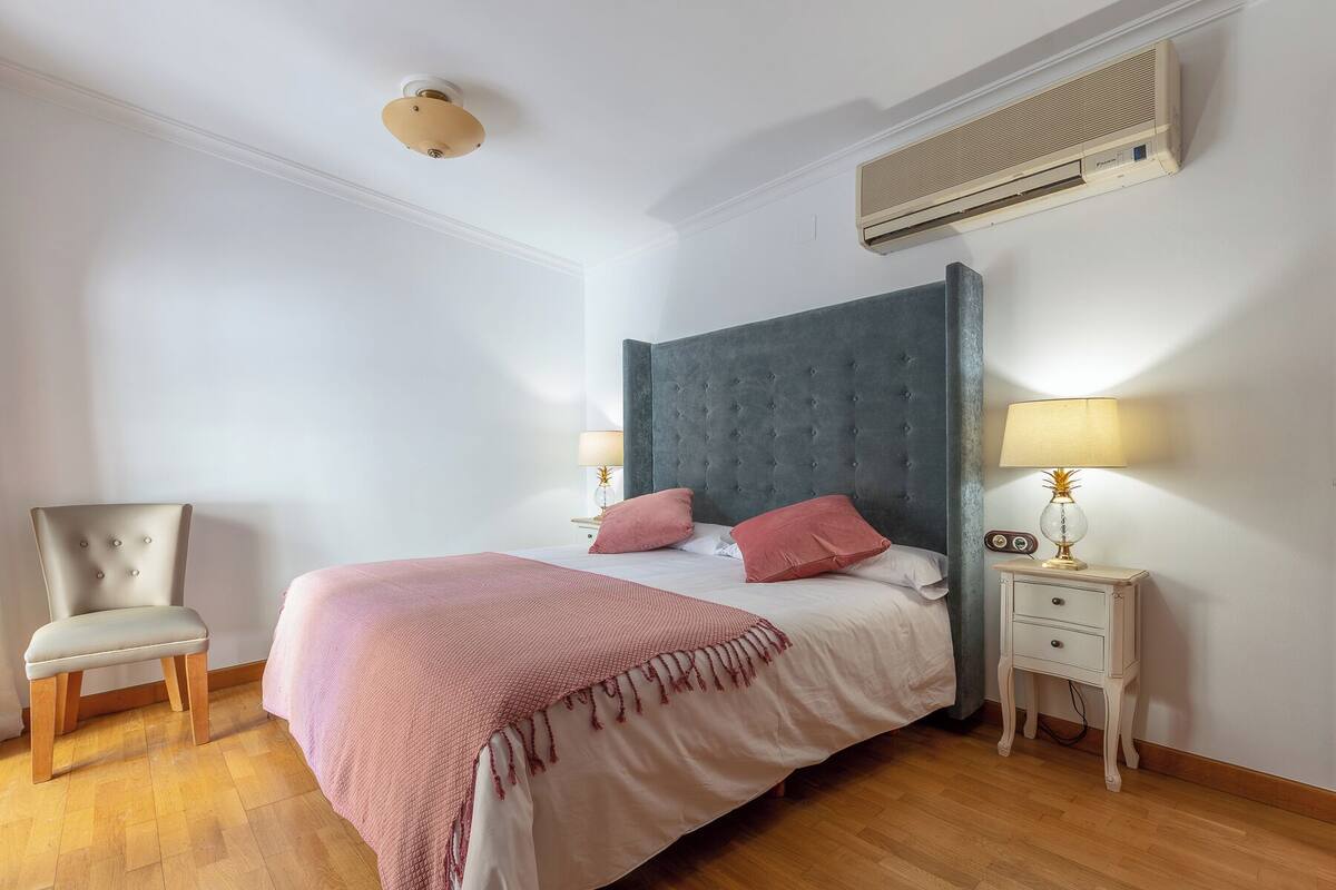 A bedroom with a bed, two bedside tables, a chair, and an air conditioning unit.