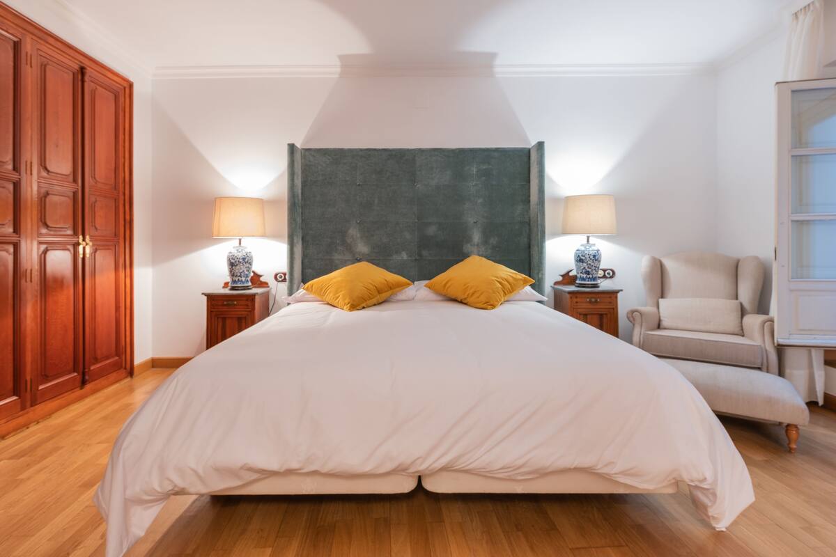 A large bed with a headboard, two bedside tables with lamps, a chair, and wooden flooring.