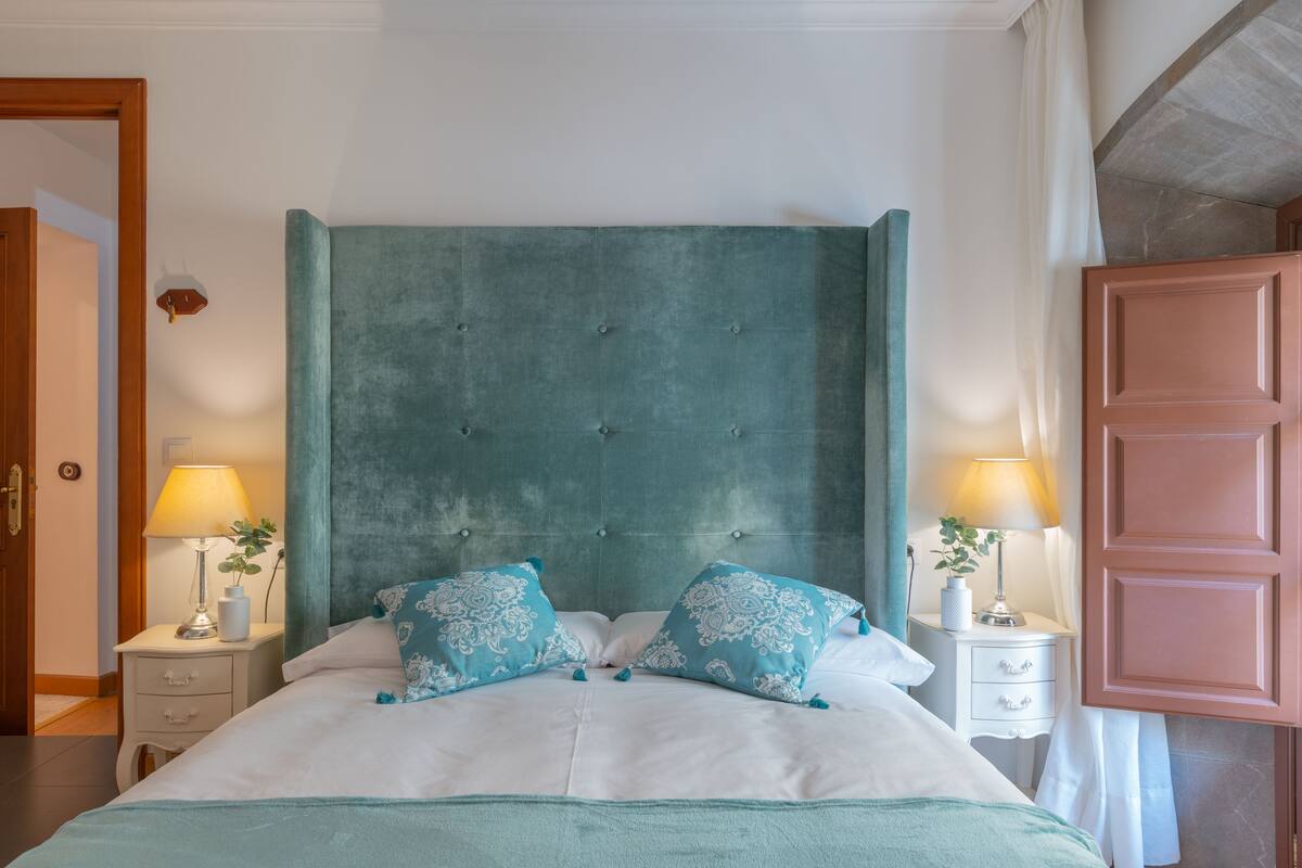 A neatly made bed with a teal headboard, two bedside tables with lamps, and a pink door.