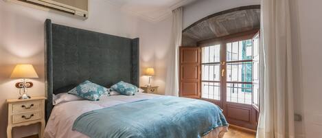 A bedroom with a large bed, two bedside tables with lamps, a window with curtains, and an air conditioning unit.