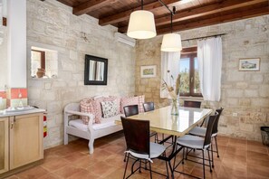 A room with a stone wall, a white sofa with floral cushions, a glass dining table with chairs, and a wooden ceiling.