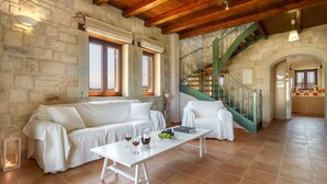 Amplia sala de estar con pared de piedra, vigas de madera en el techo, un sofá blanco, una mesa de comedor con copas de vino y una escalera con barandas verdes.