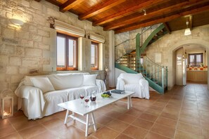 A spacious living area with a stone wall, wooden ceiling beams, a white sofa, a dining table with wine glasses, and a staircase with green railings.