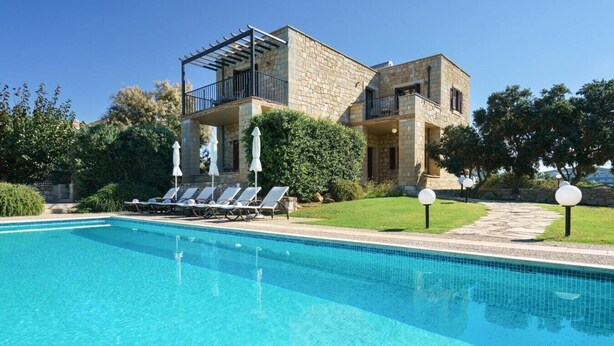 Una piscina con sillones de descanso, un edificio de piedra con balcones y un cielo despejado.