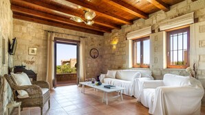 Una habitación con paredes de piedra, vigas de madera, un sofá blanco, una silla de mimbre, una mesita con vasos y vistas a través de un puerta abierta.